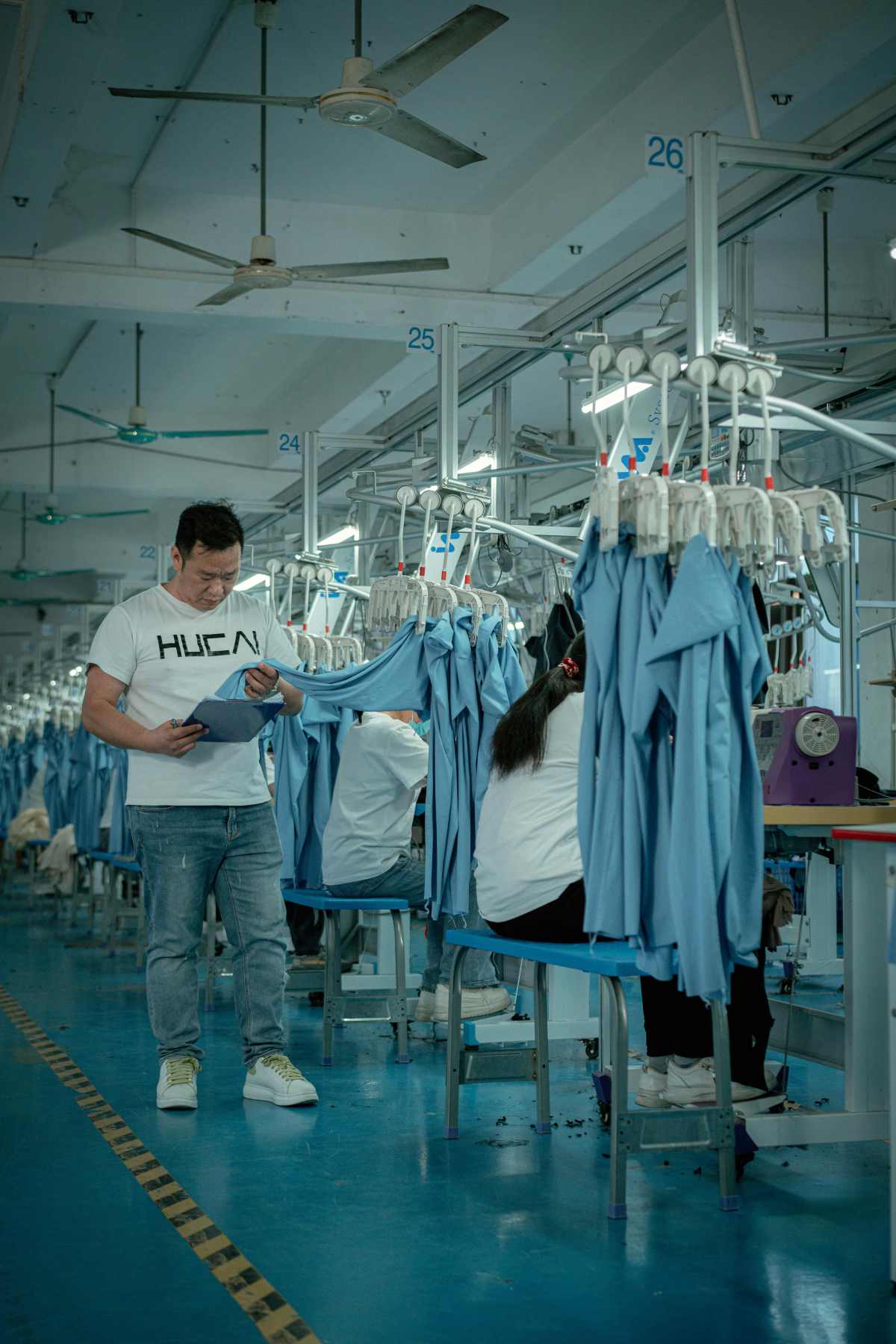 Sewing floor with numbered hanging production lines and a supervisor checking a garment against a tablet