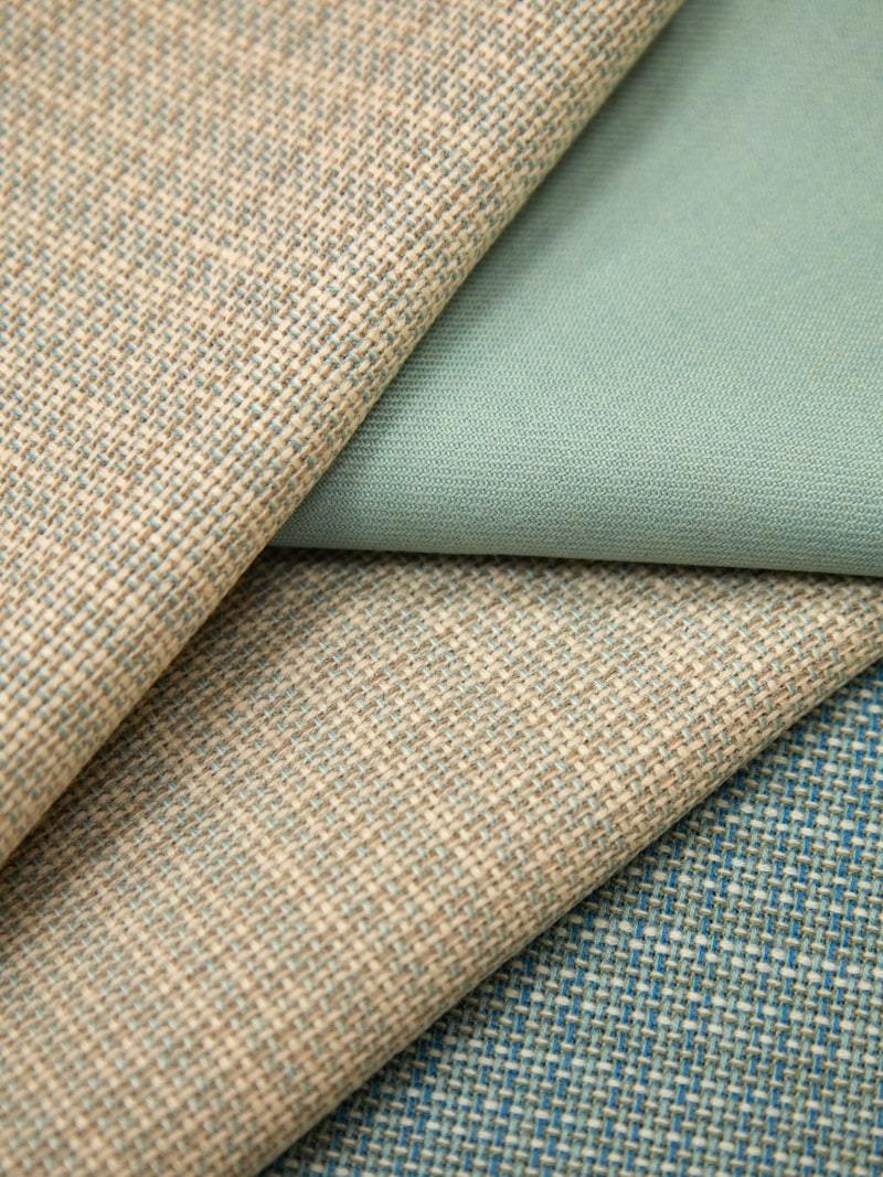 Close-up of woven fabric swatches in muted earth-tone colors including beige, sage green, navy, and tan layered together to illustrate shade and dye-lot consistency