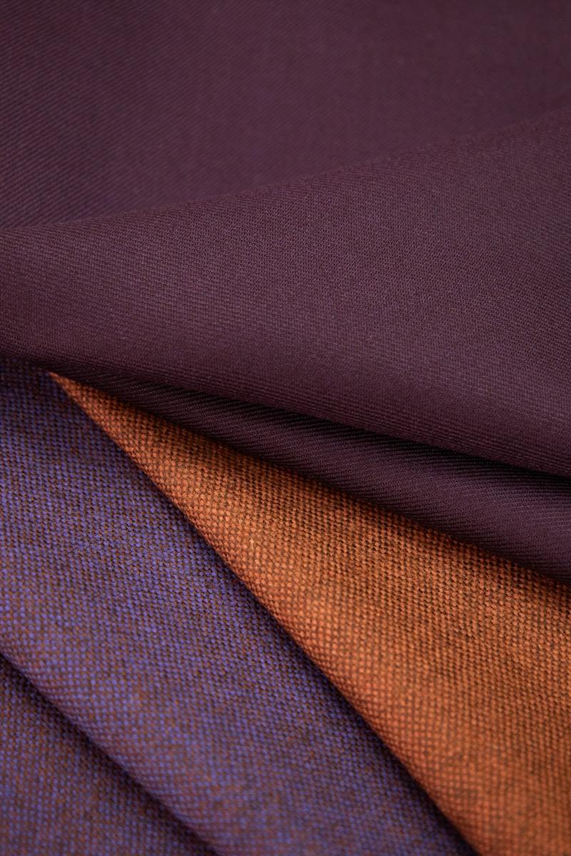 Folded stretch activewear fabric shown in three coordinated capsule colors: deep plum, burnt orange, and navy blue layered together