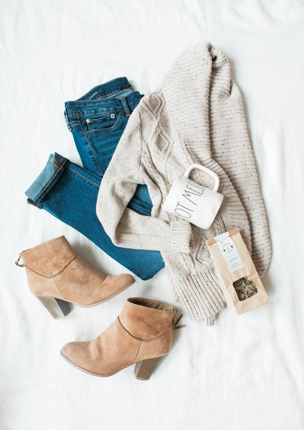 A flat-lay of a knitted sweater, jeans, suede ankle boots and a mug arranged on a white surface, styled as a winter outfit