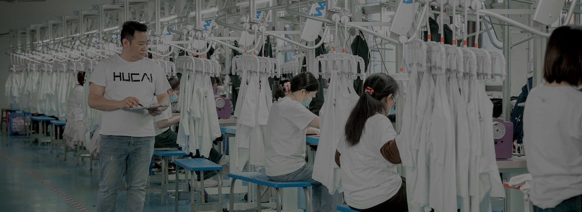 Real HUCAI garment factory hanging production line with finished garments on overhead conveyor hangers and a production supervisor reviewing a checklist, representing sample-to-bulk production control for a men's running jacket range
