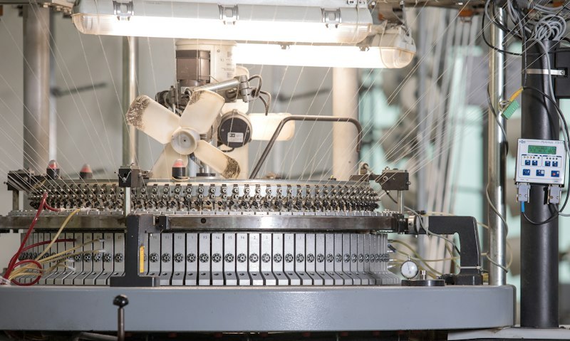 Circular knitting machine on a textile mill floor with hundreds of white yarn strands feeding into the needle bed