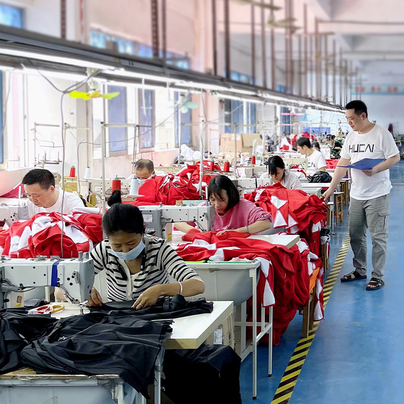 Real factory sewing floor at the HUCAI garment workshop with operators at industrial machines and a quality supervisor checking production against a clipboard alongside the line