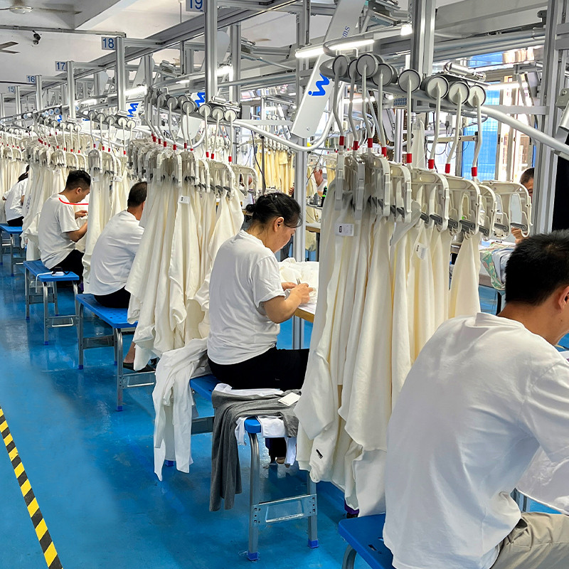 Real garment finishing and inspection area at the HUCAI workshop with finished men's training garments hanging on rails and operators recording measurements at inspection benches