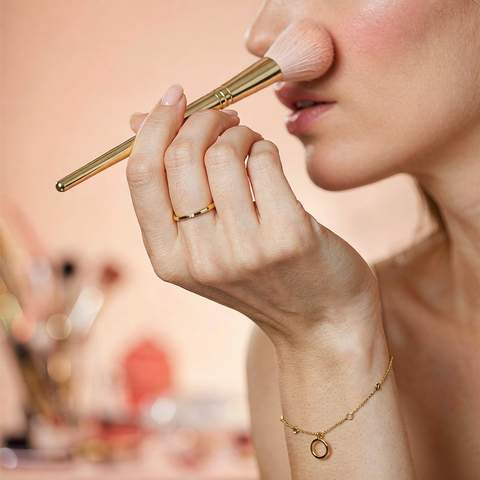 Hand wearing an 18K gold plated ring and bracelet applying makeup with a cosmetic brush, soft bokeh