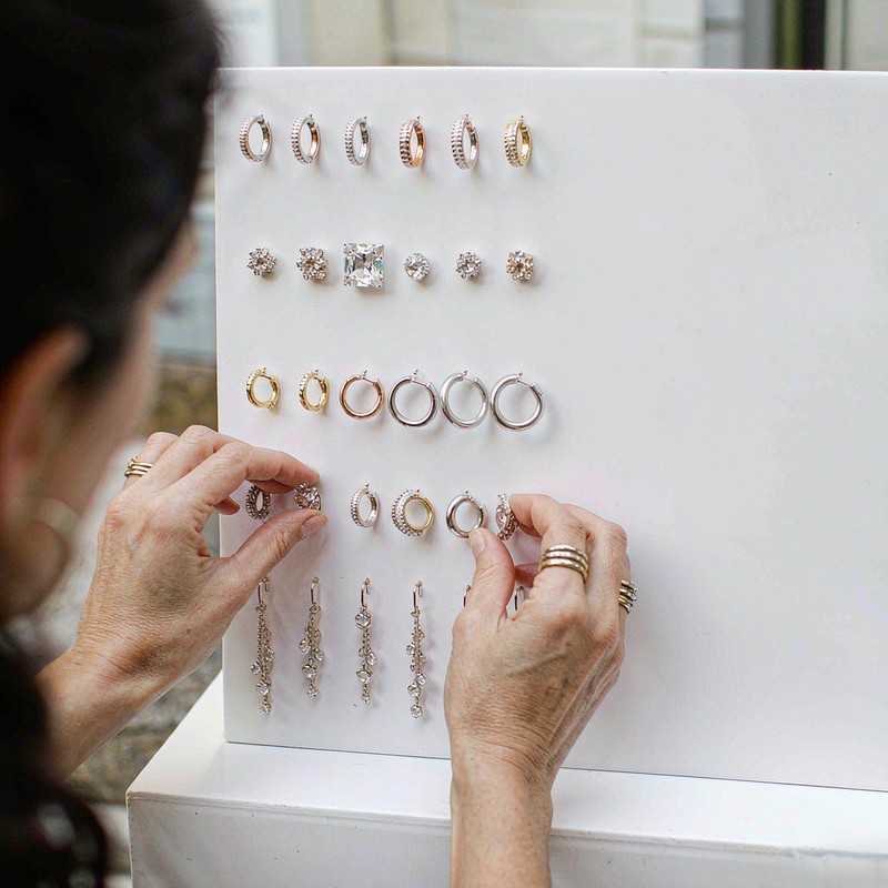 Manos de diseñadora clasificando pendientes huggie, stud, aro y gota sobre tablero de exhibición