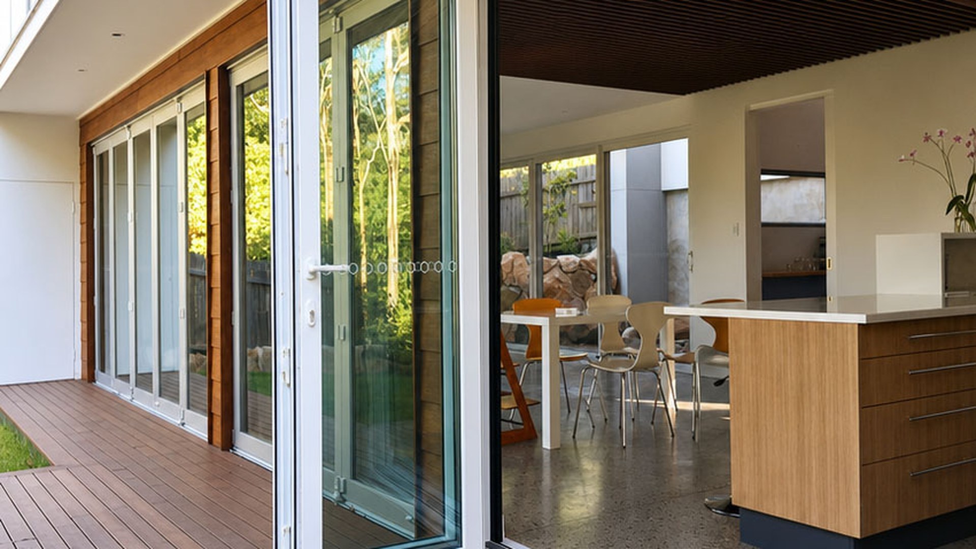 An aluminium folding door installed in a modern home, one leaf swung open along its track onto a timber deck with the living space visible behind it.