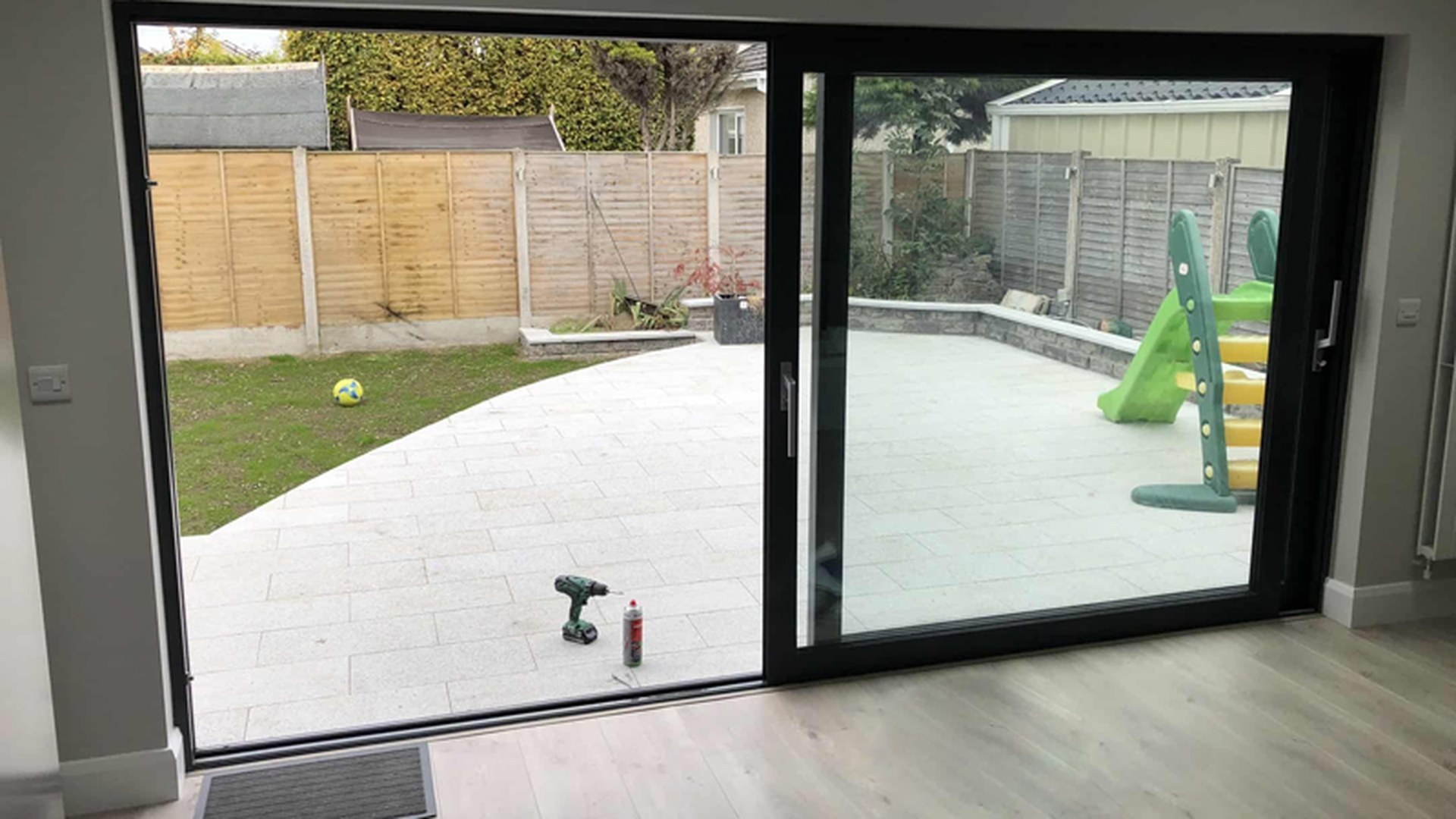 A black-framed lift and slide door photographed at installation stage, with the tools of the fitting day still on the floor and the garden beyond the glass.