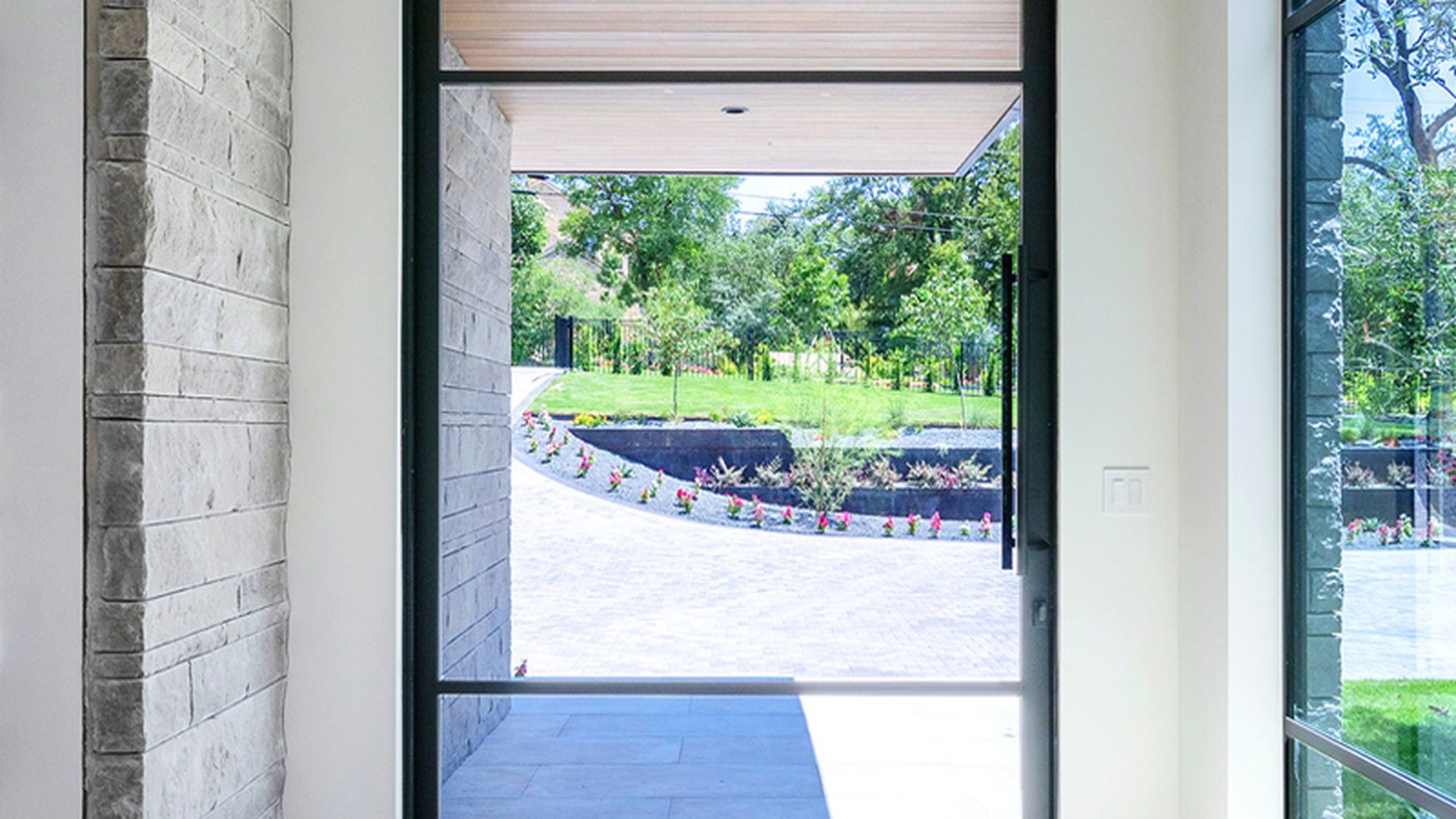 A black-framed aluminium pivot door installed in a modern living space, the leaf turning on its hidden axis across a full-height entrance that opens onto a garden.