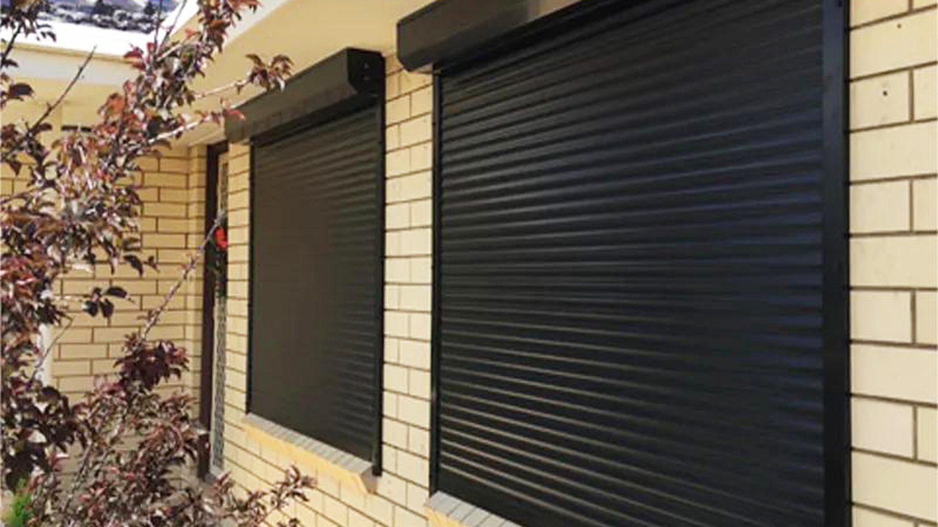 An aluminium roller shutter closed over an opening in a brick elevation: curtain, guides and head box are one system, and once it is down the opening is a barrier rather than a shade.