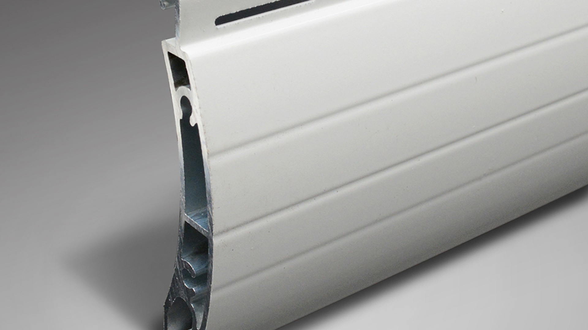 The cut end of an extruded aluminium shutter slat, showing the hollow chambers that stiffen the curtain and the interlock that keeps one slat engaged with the next.