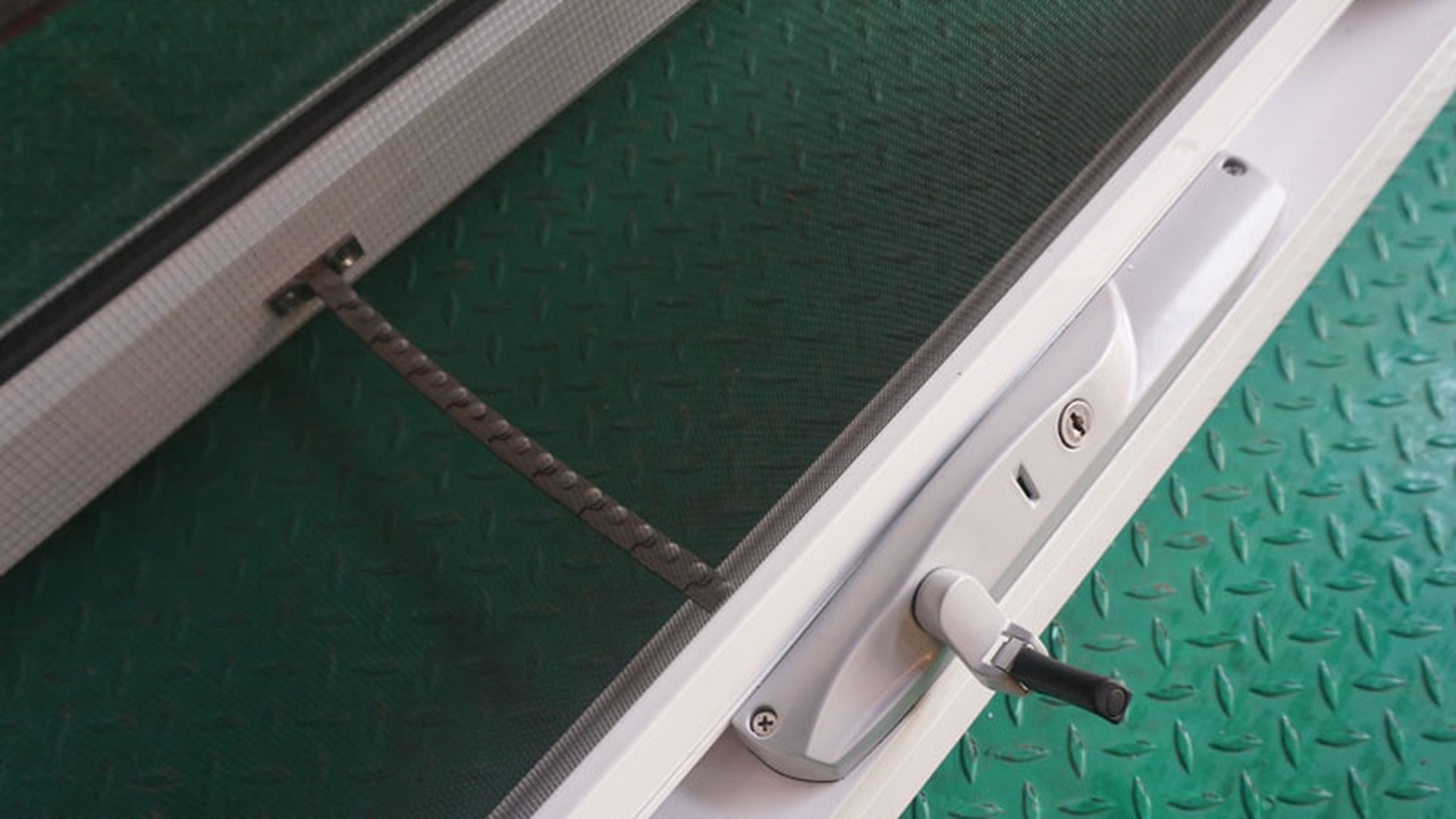 A chain winder and its lock, photographed on a finished white frame with the flyscreen in place: the operator, the screen and the lock all share the same narrow bottom rail.