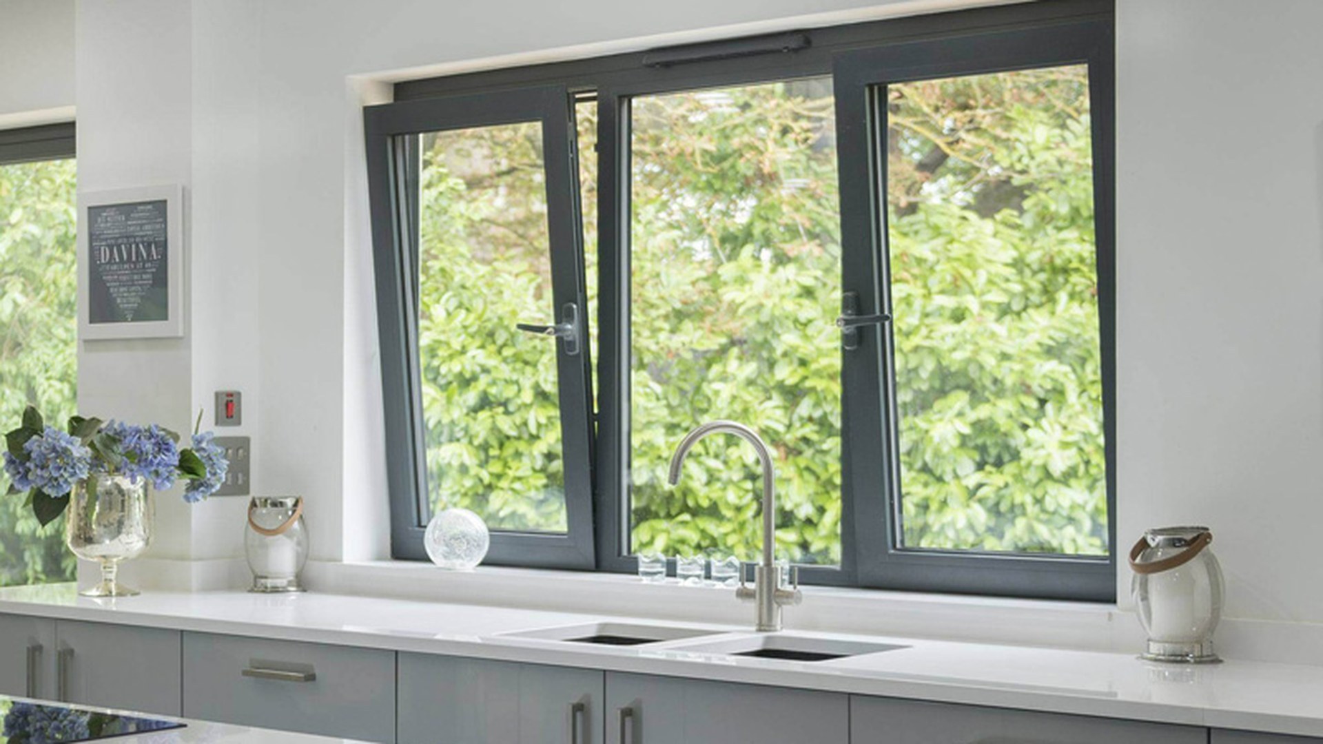 Tilt sashes over a kitchen sink run in charcoal frames: the sash leans into the room at the top, so the vent gap sits at working height where nobody has to reach over the tap to open it.