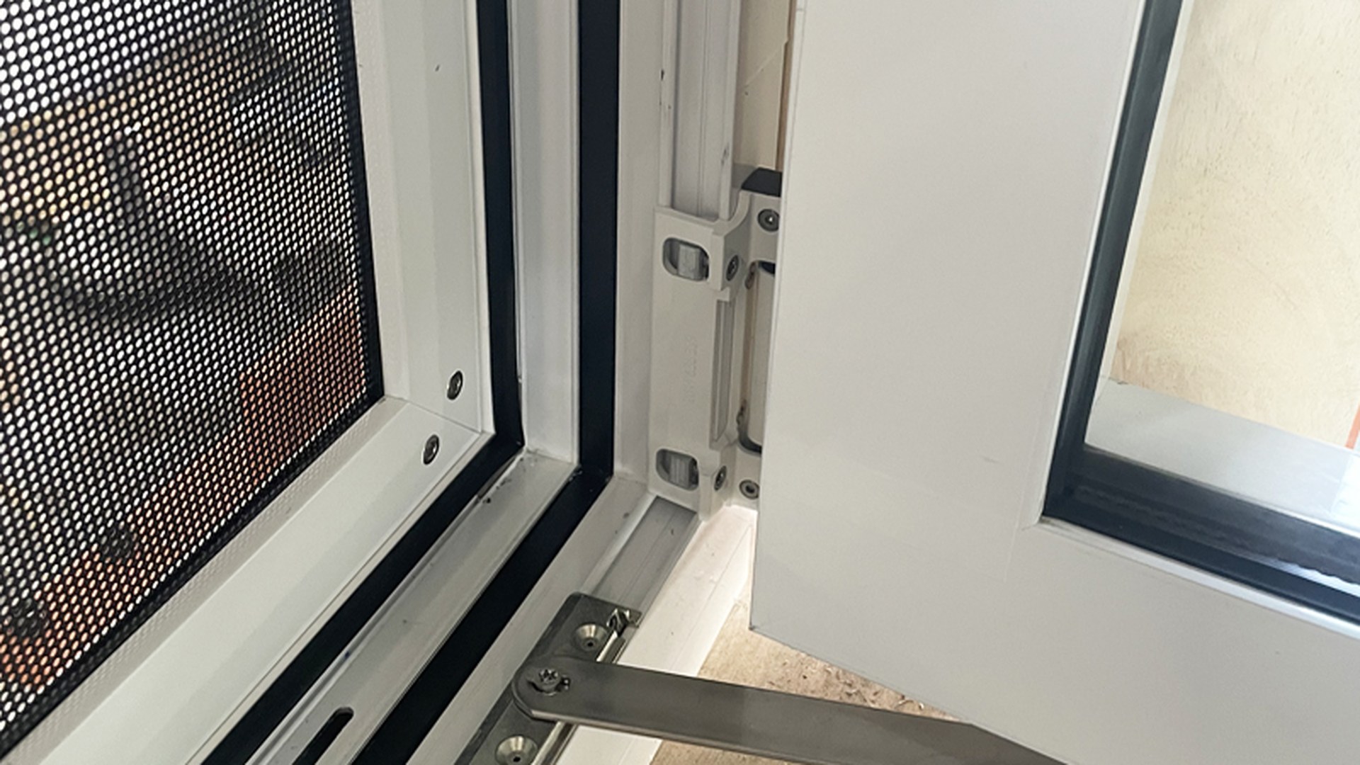 The hinge side and the limit arm at the bottom corner of an installed white sash, with the flyscreen carried in its own frame: this is where a tilt and turn window is loaded every time it is operated.