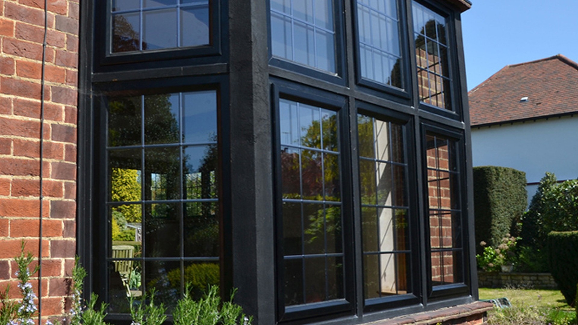 A 45-degree bay in black aluminium with Georgian bars: three faces of glass pushed beyond the wall line, with the roof and the joint at each angle doing the work that a flat window never has to do.