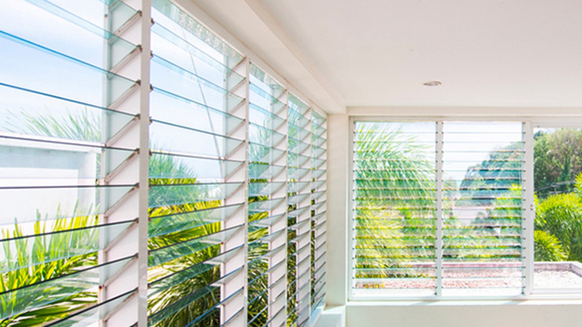 Three louver panels turning a corner with the blades open: with no sash to swing, the same blade stack ventilates a glazed corner without taking floor space.