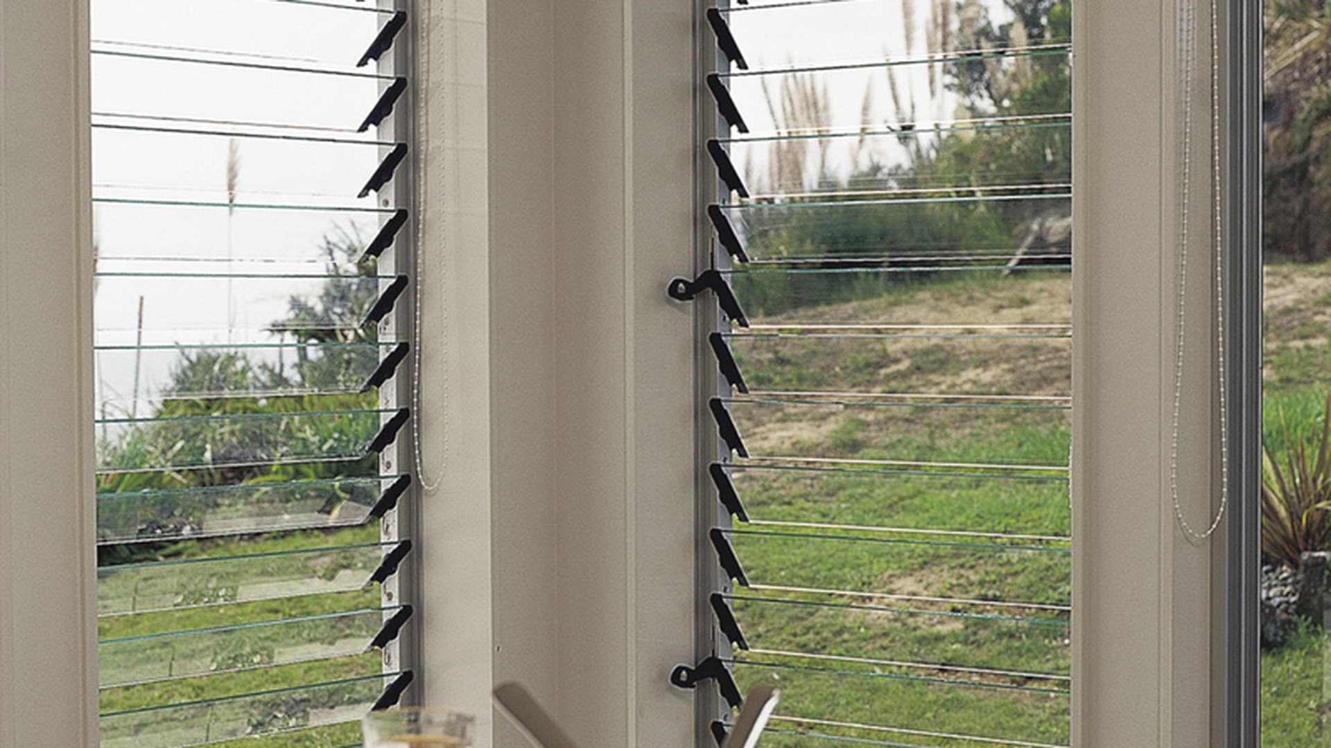 Two adjustable louver panels beside a dining table: the chain on the jamb sets the blade angle, so ventilation is tuned without opening anything into the room.