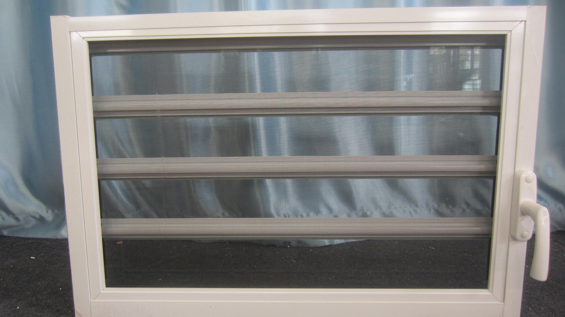 A complete louver unit on the factory stand: blades, jamb, carrier and fixed flyscreen arrive as one frame, so the screen plane has to be decided before the profile is cut.