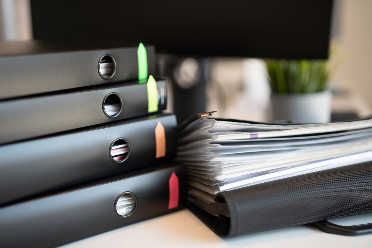 Ring binders filled with documents on an office desk beside a monitor, representing a per-product technical file