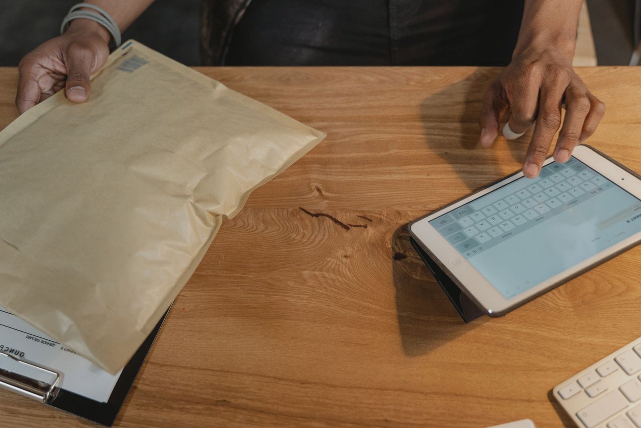 Person holding a padded parcel in one hand while using a tablet to review an online order on a wooden desk