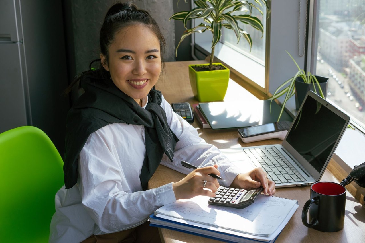 A young sourcing manager at her desk working through supplier cost sheets with a calculator, printed documents and a laptop in a bright office, the starting point of a desk-based digital factory audit.