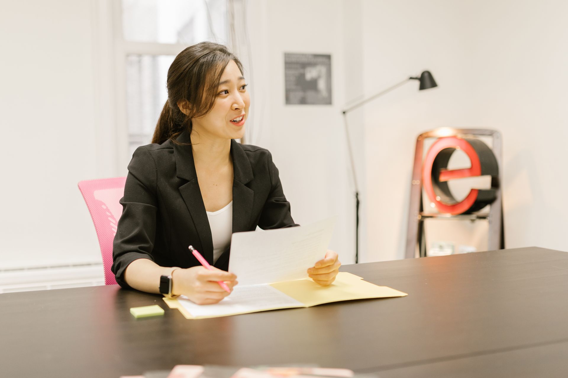A young sourcing analyst in a black blazer reviewing printed supplier quotations with a pen at a bright office desk.