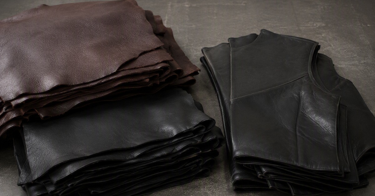 Stacks of dark brown and black leather hides beside cut jacket panels on a factory bench