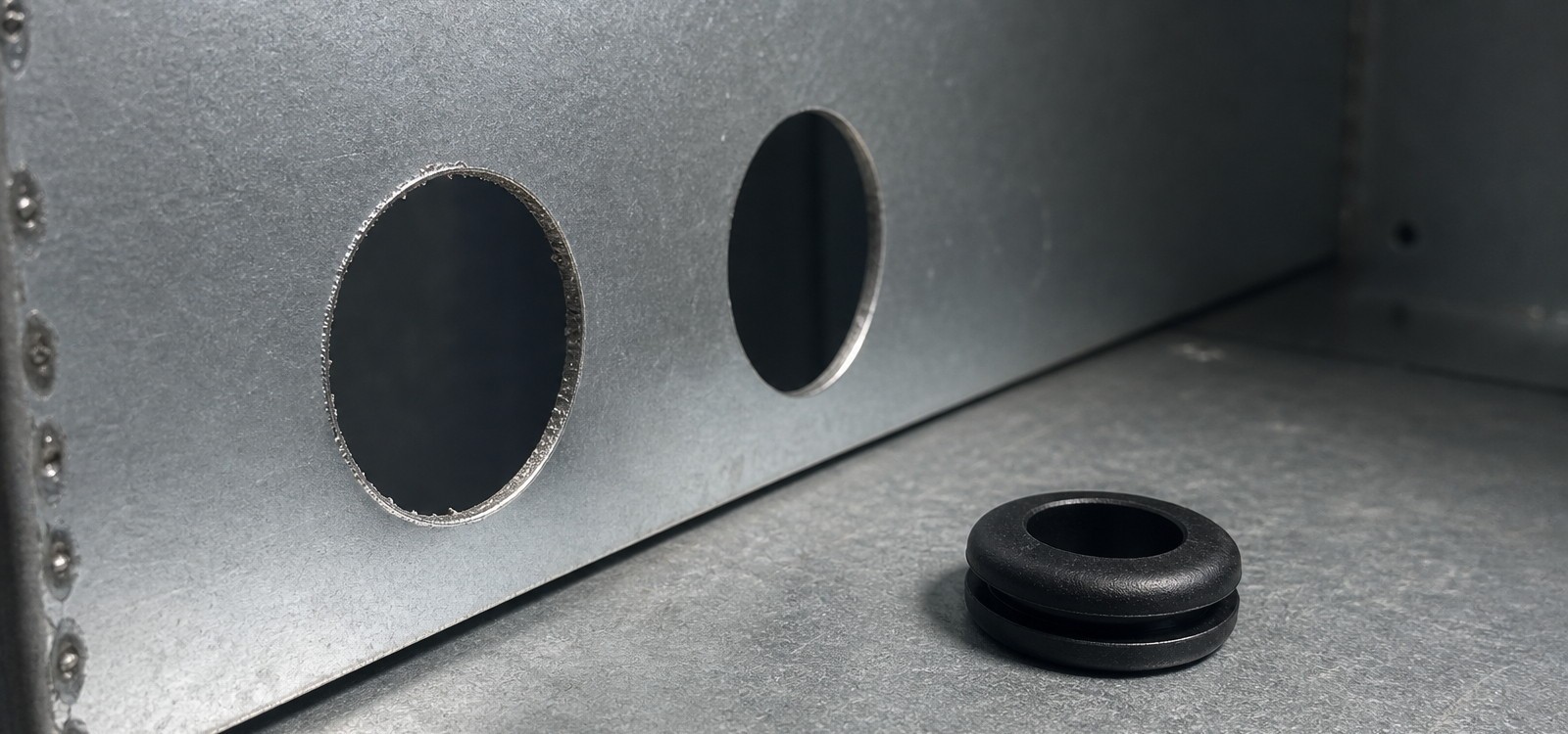 Thin galvanised sheet metal panel with stamped round holes showing sharp cut edge and burr next to a rubber grommet