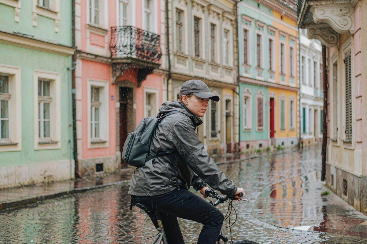 Commuter in a matte charcoal waterproof jacket cycling through a European old-town street in light rain