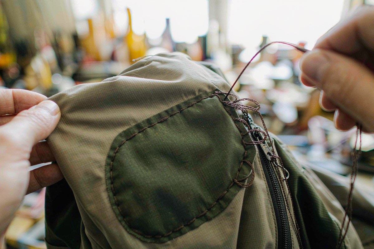 Hands stitching a fabric repair patch onto the sleeve of a muted green technical shell jacket