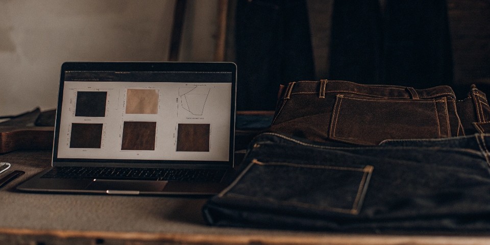 A workspace showing a laptop with an online store, design sketches, denim swatches, and a pair of finished jeans.