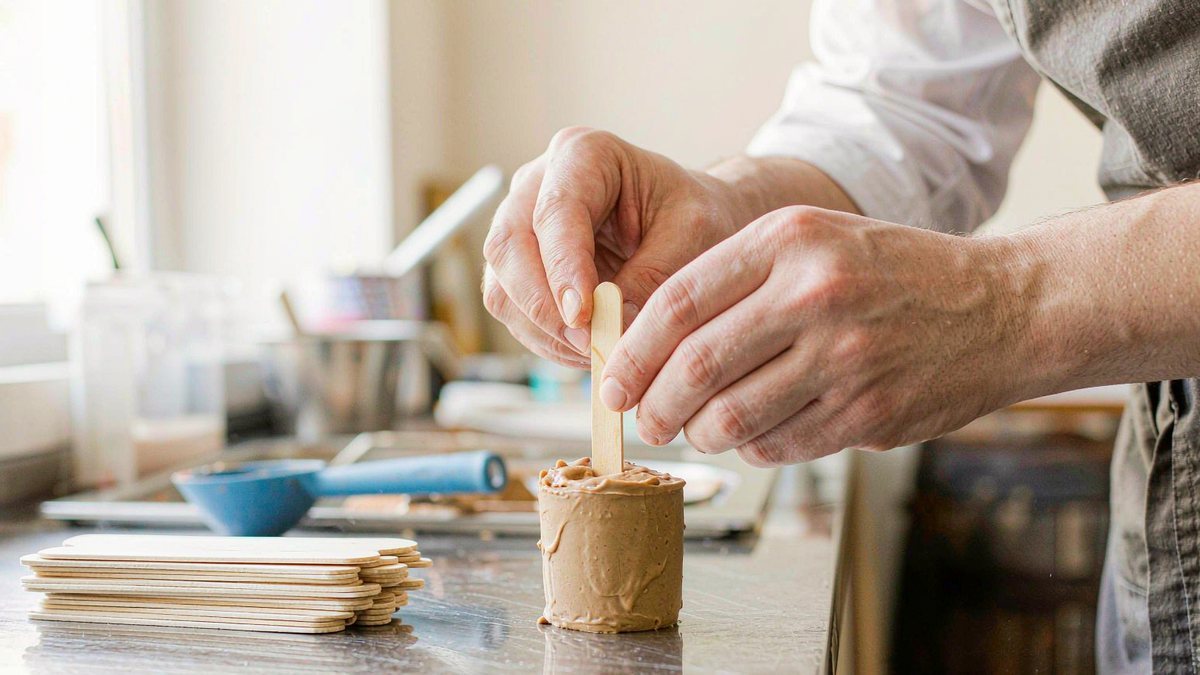 Hands inserting a wooden stick into a freshly filled ice cream mould in a small-batch workshop