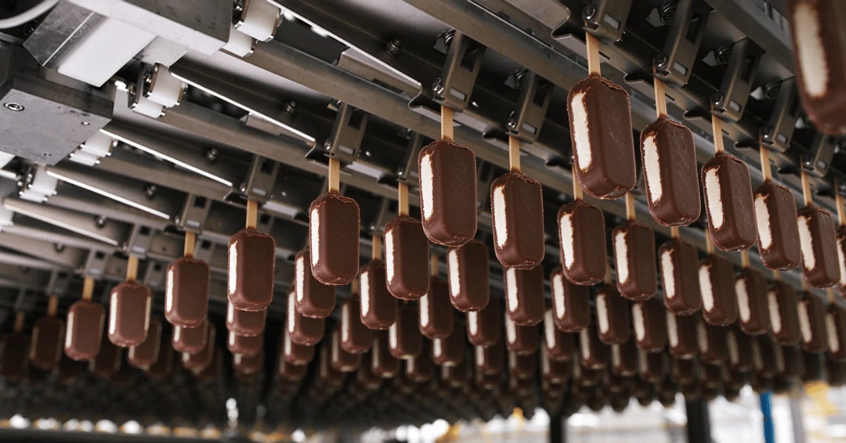 Chocolate-coated ice cream bars hanging from wooden sticks moving through a high-speed moulding and coating line