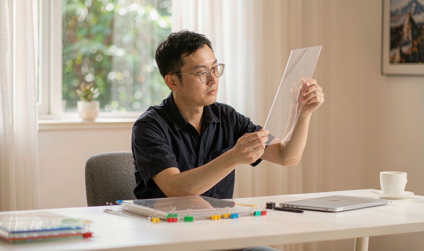 A design engineer holding a transparent polycarbonate overlay film up to the light, with PET and PMMA sample sheets arranged on the desk