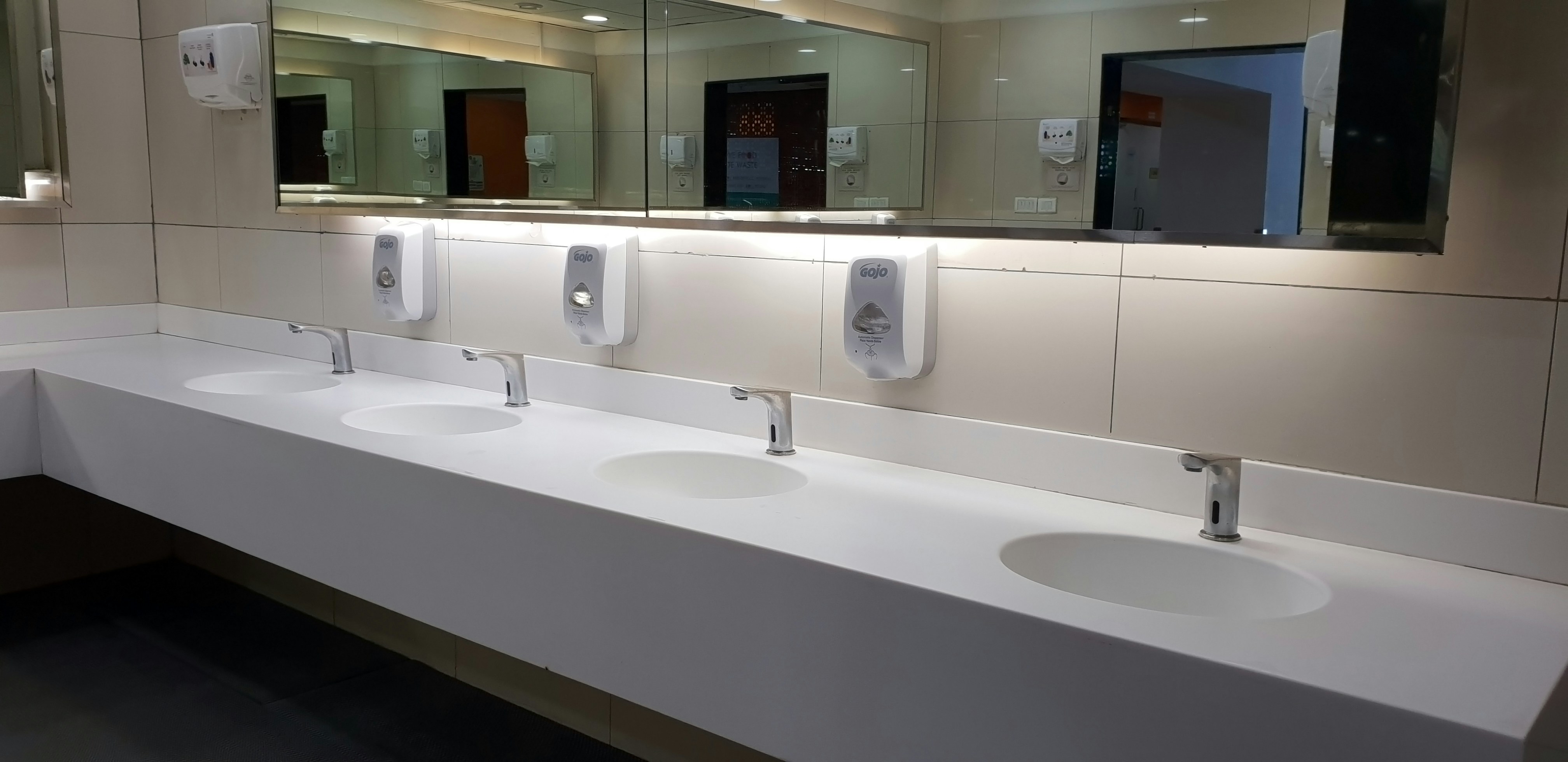 A modern public restroom featuring a long, white solid surface trough sink with multiple wall-mounted faucets.