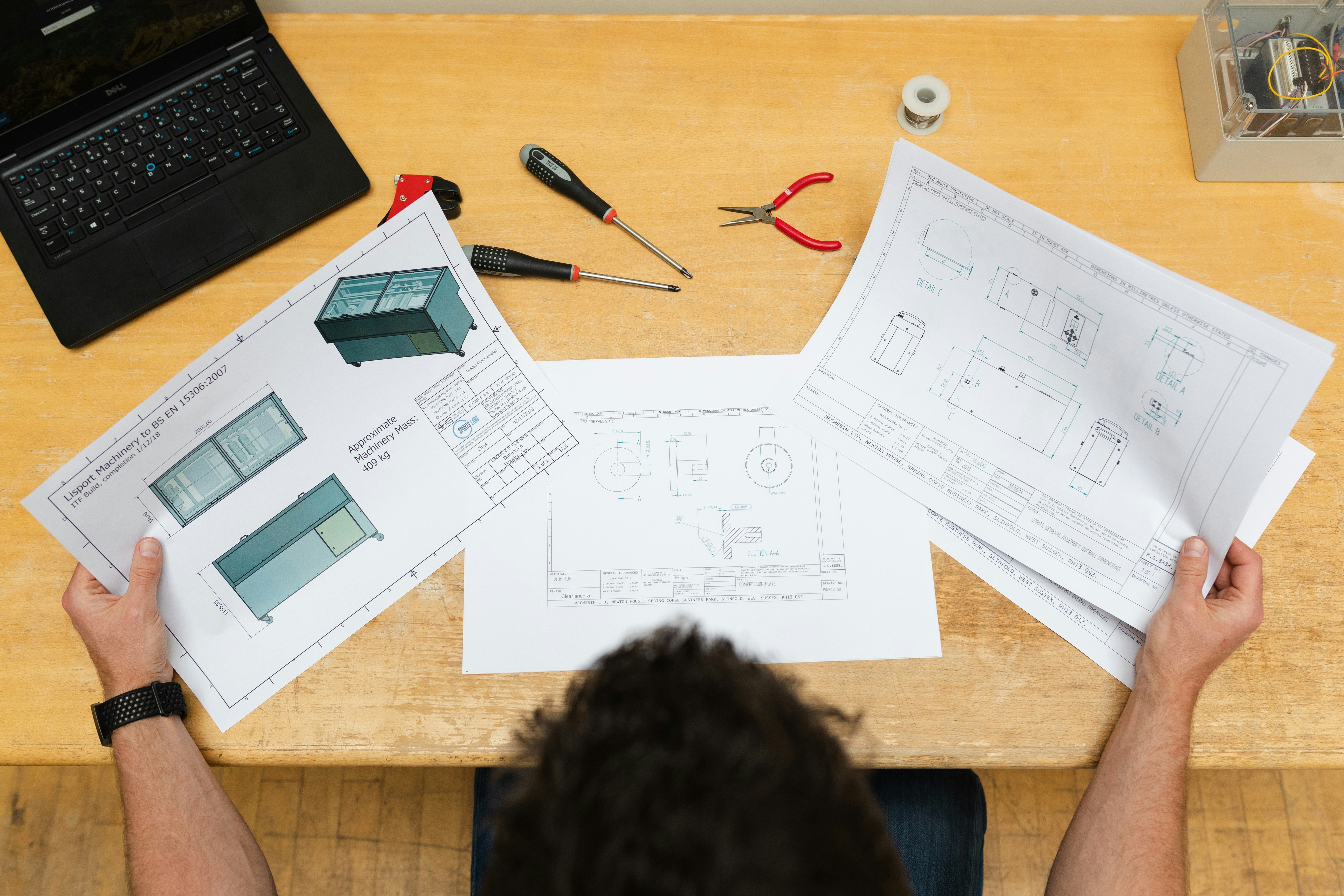 A person at a desk with blueprints, a calculator, and a laptop showing a spreadsheet for project budgeting.