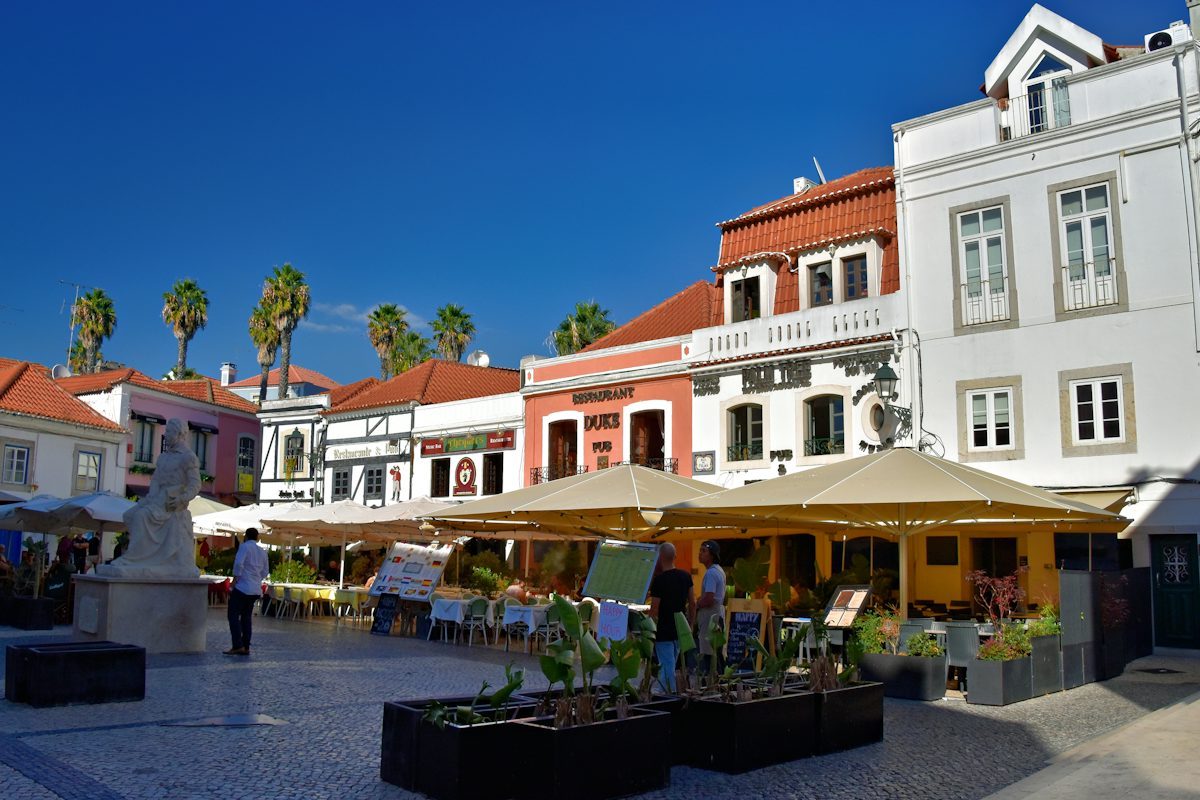 Outdoor restaurant tables under large umbrellas with palm trees behind