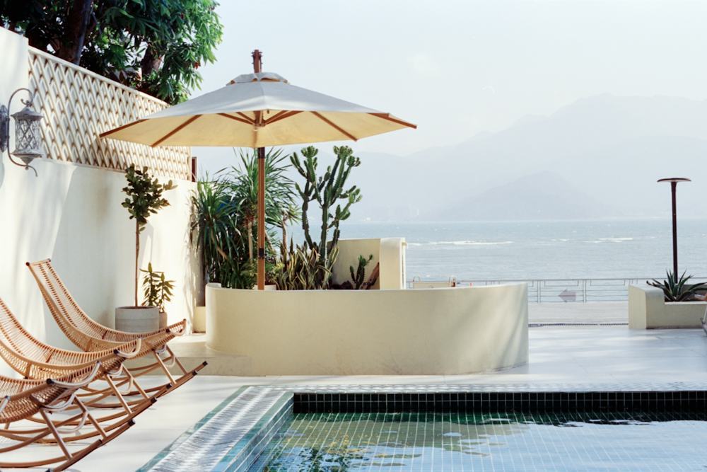 Poolside terrace with an open cream umbrella, rattan chairs and a hazy sea view