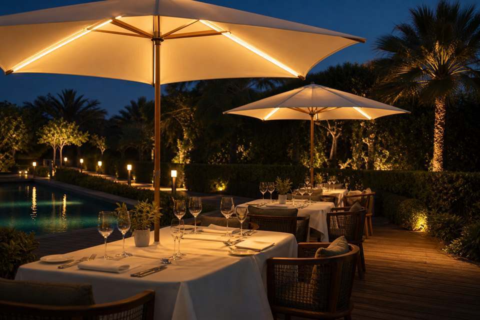 Center pole umbrellas lit from inside above set tables on a garden dining terrace at dusk
