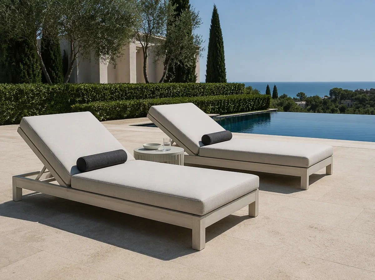 Two pale outdoor lounge daybeds with cream cushions on a resort pool deck beside still blue water
