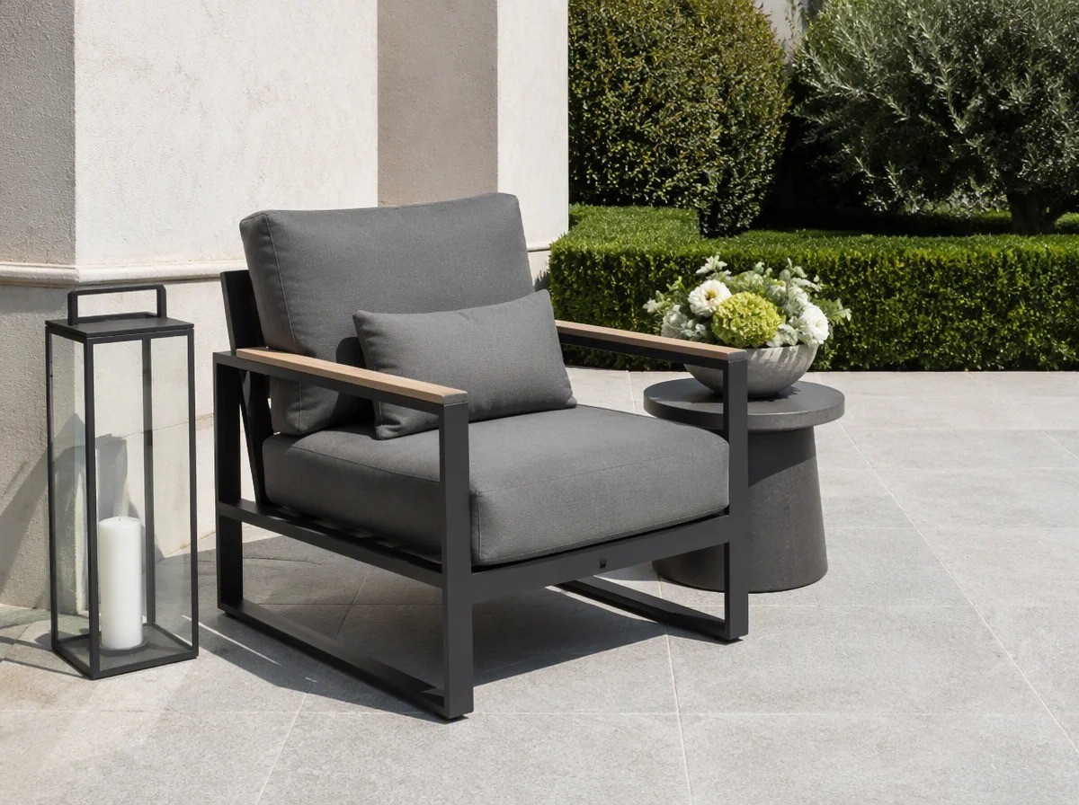 Outdoor lounge sofa with solid teak armrests and grey cushions beside a glass hurricane lantern and a bowl of cut flowers on a stone terrace