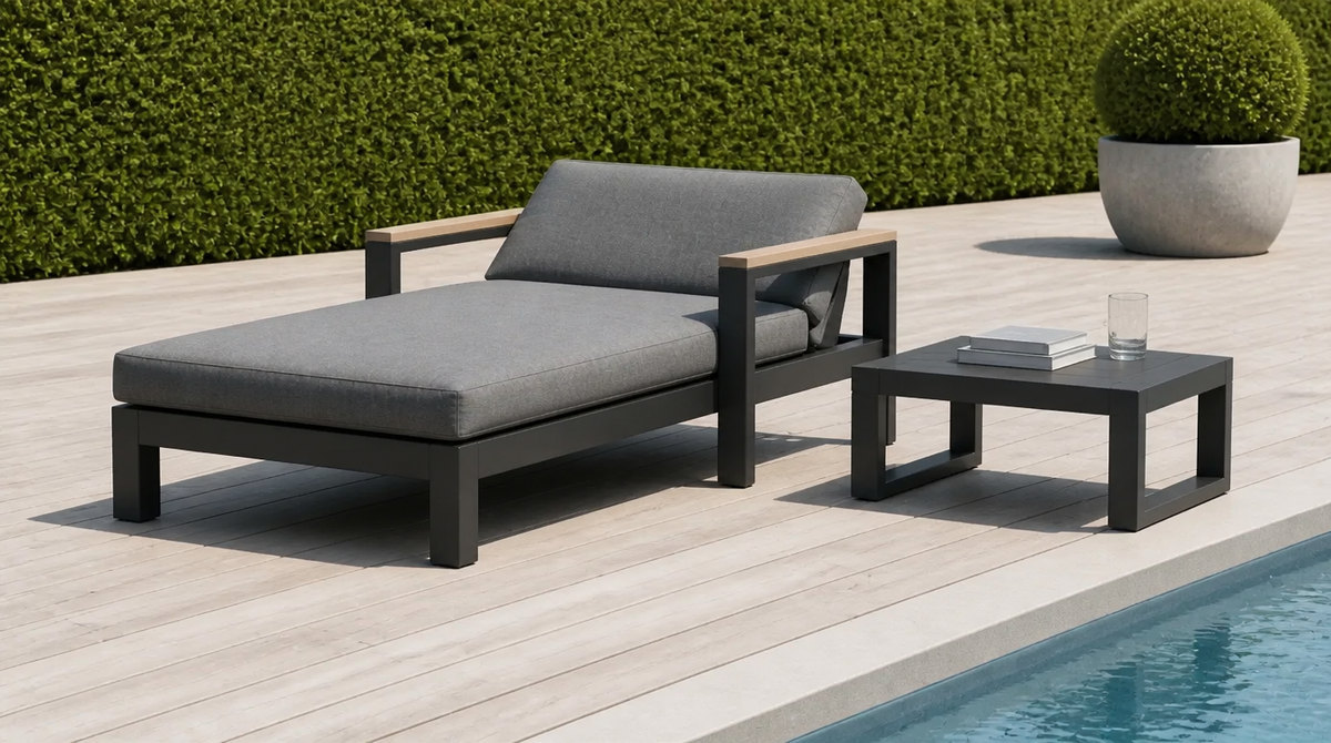 Outdoor daybed with a grey quick-dry cushion on a poolside timber deck beside calm blue water, with a low side table holding books and a glass