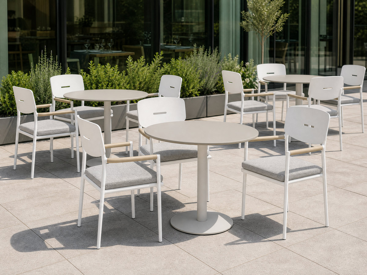 Restaurant terrace laid up for service with several identical white aluminium armchairs around round tables beside a glazed frontage