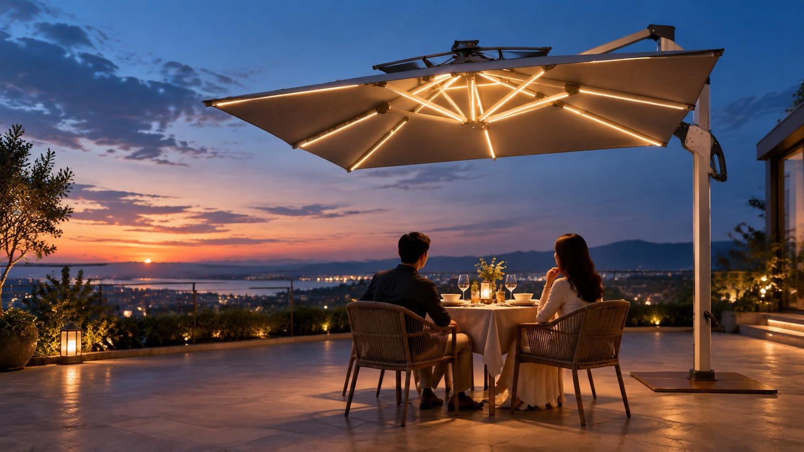 A lit cantilever patio umbrella over a candlelit table for two on a hotel terrace at dusk, with the sea and city lights beyond