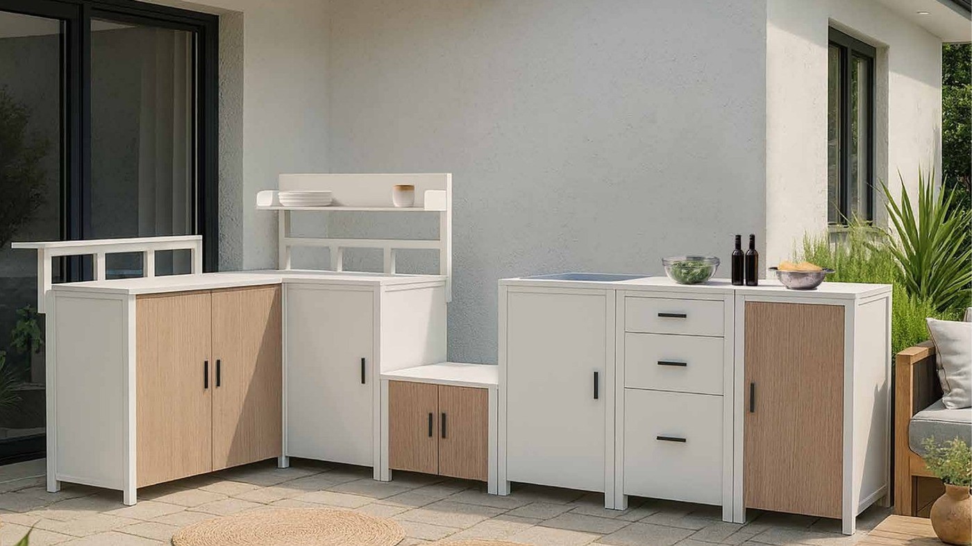 A white aluminium modular outdoor kitchen with light wood-look fronts built against a house on a paved terrace, with jute rugs and a dining table