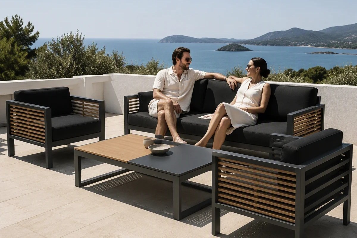 Dark-framed outdoor sofa and armchairs with wood-slat detail on a terrace behind a parapet wall overlooking a bay