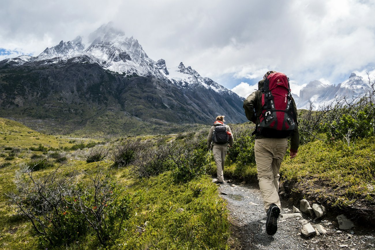 Two hikers on a mountain trail toward snow-capped peaks