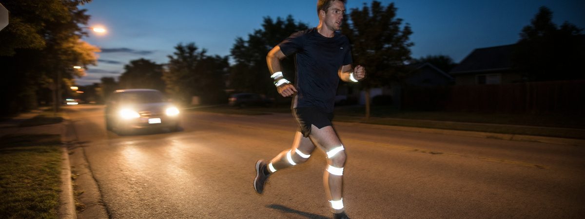Caucasian European-American male runner on a dusk road with retroreflective strips on the ankles, knees and wrists of his running shorts and sleeves glowing in car headlight beams