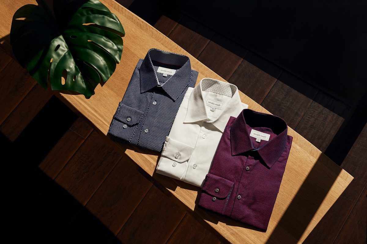 Three folded dress shirts in navy, white and burgundy on wooden bench