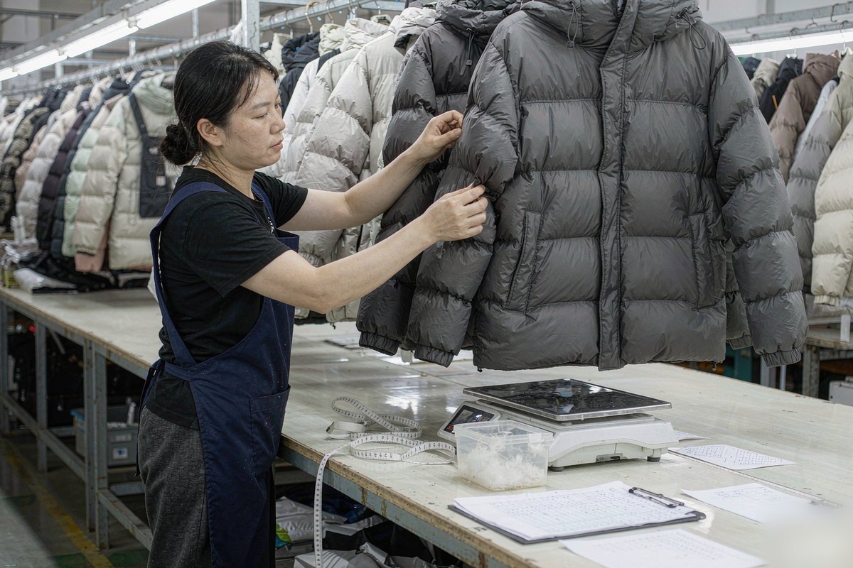 Quality control inspector in a garment factory comparing a finished charcoal gray quilted puffer jacket against a sealed approved sample beside a scale, tape measure and measurement chart